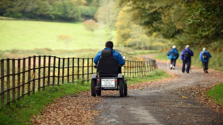 Someone riding a mobility scooter on a tarmacked path, in an autumnal woodland.